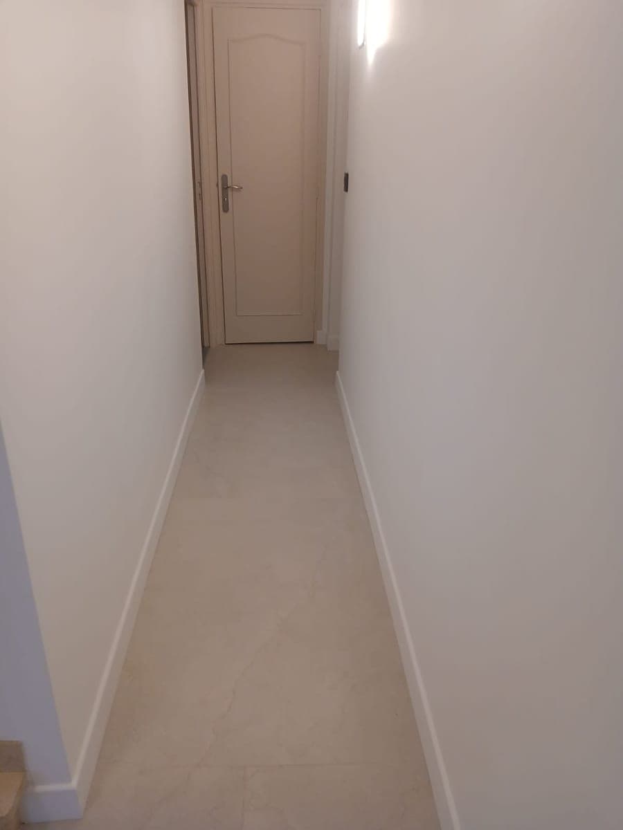 Couloir étroit au carrelage clair et aux plinthes blanches, porte au fond